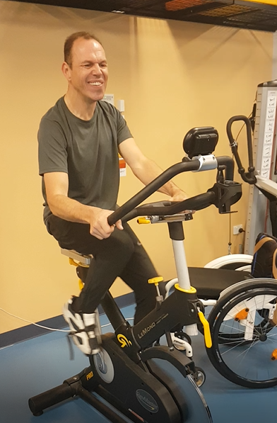 Thomas cycling during his session at NeuroMoves in Lismore. His wheelchair can be seen in the background. 
