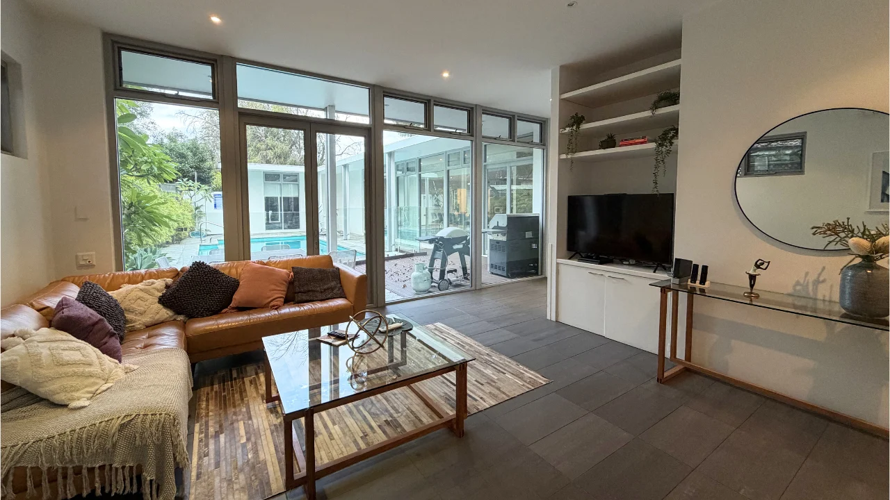 Living room at SCIA Transition House, with a lounge on the left and a wall-mounted TV on the right. Floor-to-ceiling glass doors at the back open onto an outdoor pool and BBQ area.