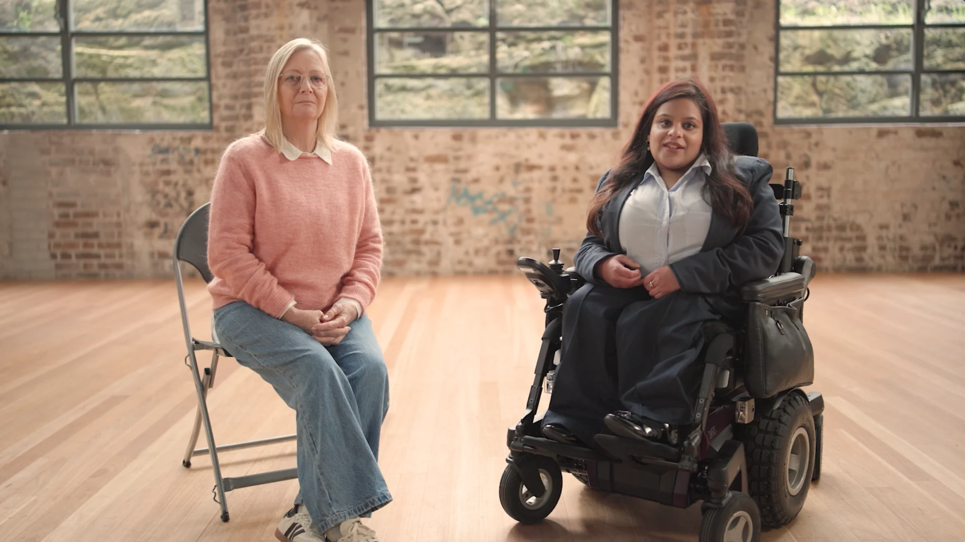 Two people sit side by side in a bright studio space with exposed brick walls and large windows, one seated on a chair and the other using a powered wheelchair, facing the camera.