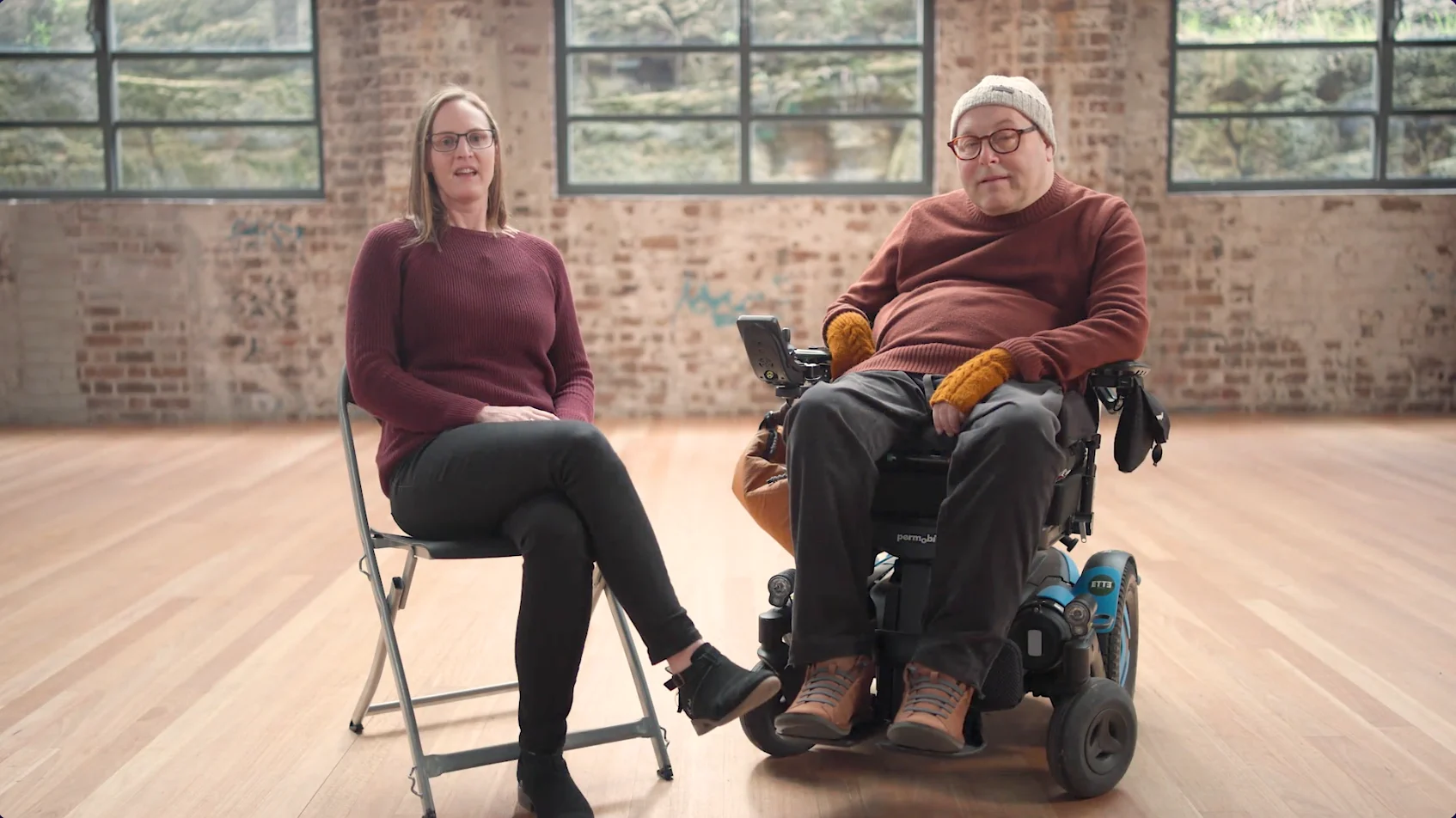 Two people sit side by side in a bright studio space with exposed brick walls and large windows, one seated on a chair and the other using a powered wheelchair, facing the camera.