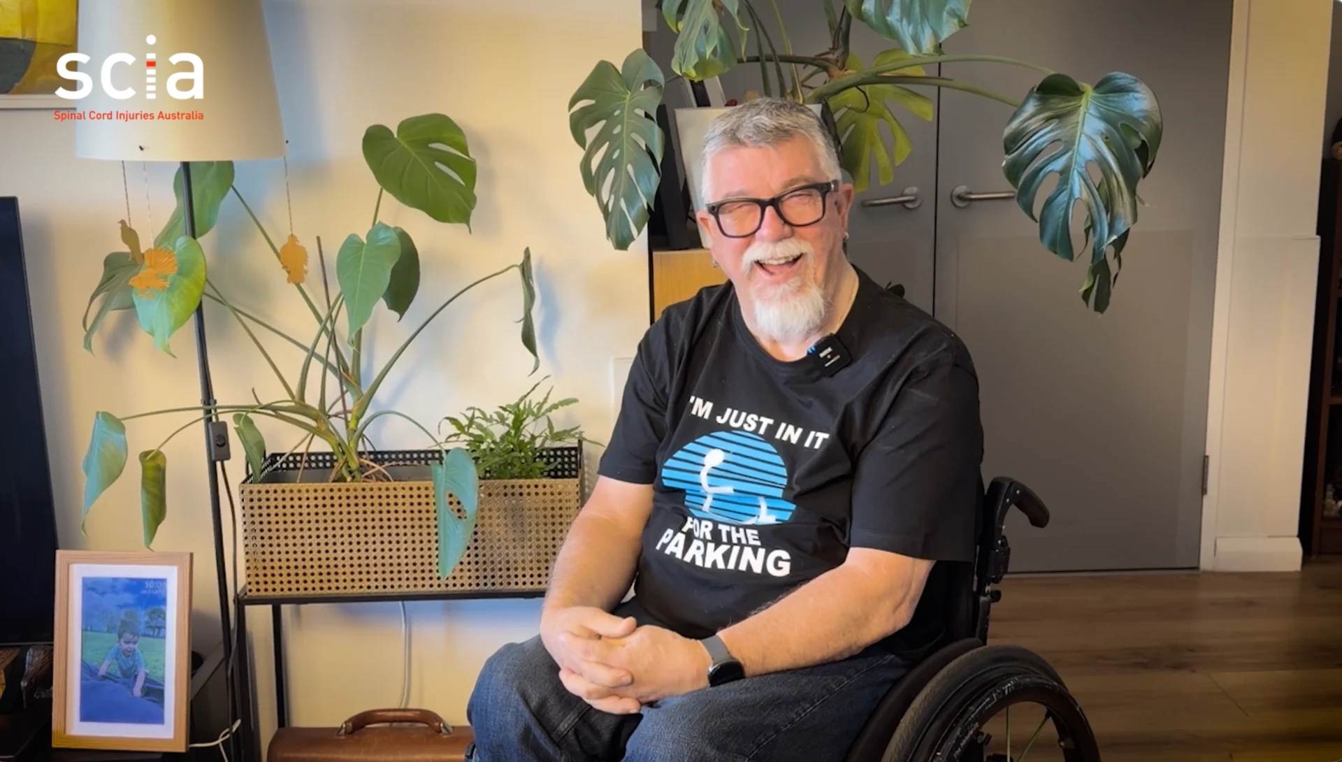 Stuart is laughing, seated in his wheelchair in a living room setting with plants behind him.