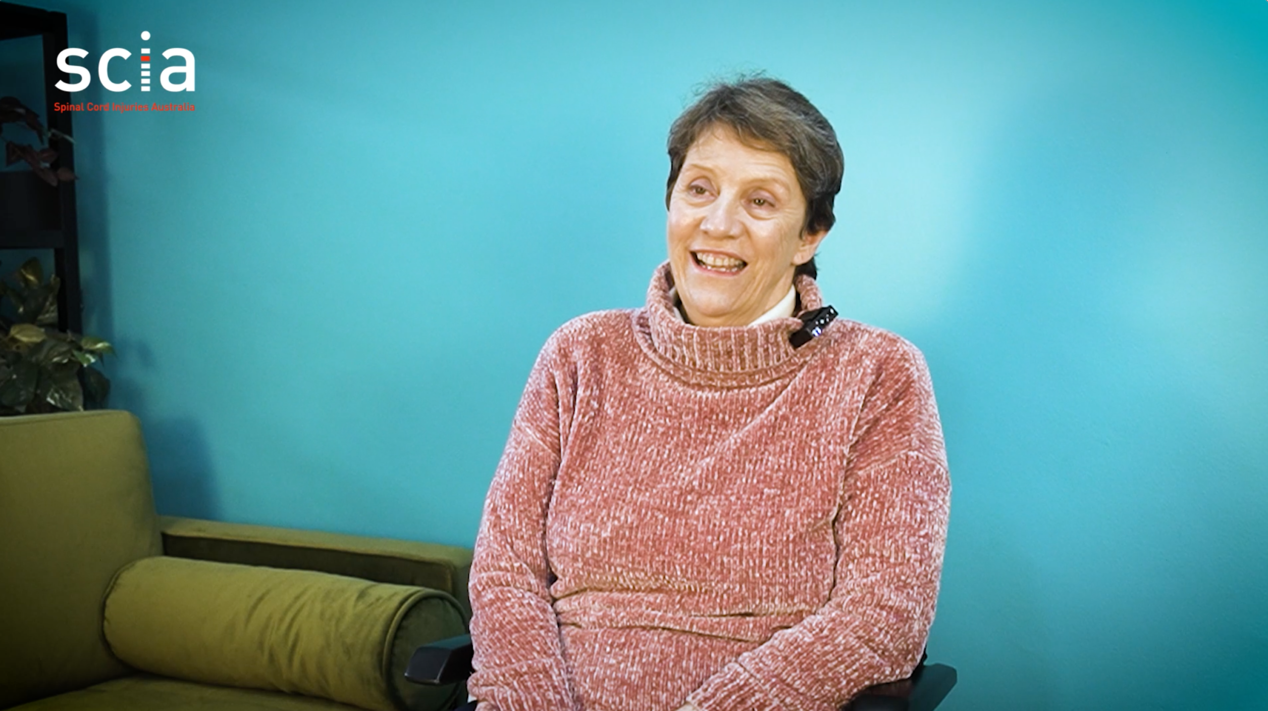 Anne is smiling and seated next to a green lounge against a teal wall.